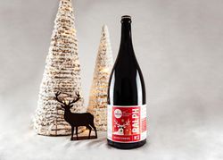 KI generiert: Das Bild zeigt eine Flasche Bier mit weihnachtlichem Etikett neben einem kleinen Hirsch-Dekor und zwei schneebedeckten, beleuchteten Weihnachtsbaum-Dekorationen. Der Hauptinhalt ist die festliche Präsentation der Bierflasche.
