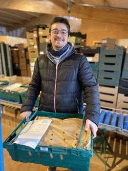 KI generiert: Ein Mann hält eine grüne Kiste mit verpackten Waren in einem Lager. Im Hintergrund sind Regale mit weiteren Kartons und Kisten.