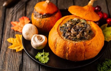 KI generiert: Das Bild zeigt zwei ausgehöhlte Kürbisse, die mit einer herzhaften Füllung, wahrscheinlich bestehenden aus Pilzen und Gemüse, gefüllt sind. Sie sind auf einem Teller angerichtet, umgeben von Pilzen, Herbstblättern und Beeren auf einem Holztisch.