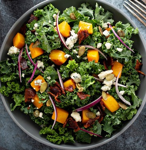 KI generiert: Ein Salat aus Grünkohl, Kürbis, roten Zwiebeln, Blauschimmelkäse, getrockneten Tomaten und Kürbiskernen in einer Schüssel. Der Salat sieht frisch und bunt aus.